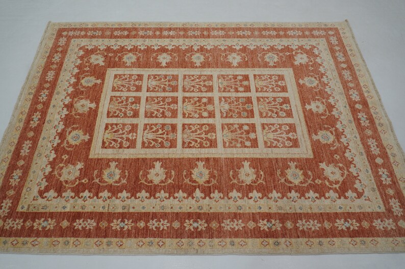 May include: A traditional rug with a red background and a beige pattern. The rug has a central design of a grid of squares with floral motifs. The border of the rug is decorated with a repeating pattern of small flowers.