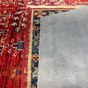 May include: Close-up of a red oriental rug with intricate patterns in blue, yellow, and orange. The rug's border features a detailed design. A gray, textured backing is partially visible, providing a non-slip surface.