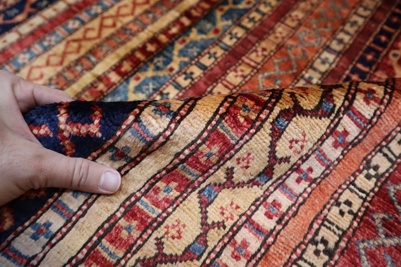 May include: A close-up of a colorful rug with intricate patterns in shades of red, blue, and cream. The rug is woven with a fine texture and has a traditional design.