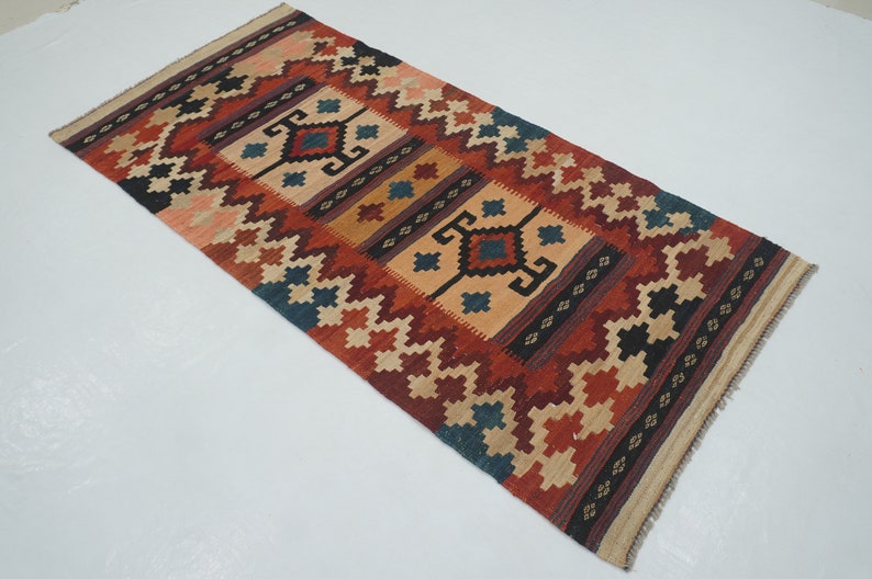 Puede incluir: Alfombra rectangular con un dise&ntilde;o geom&eacute;trico en tonos rojos, beige, azul y negro. La alfombra presenta un dise&ntilde;o central con formas de diamante y un borde con patrones repetidos. La alfombra est&aacute; probablemente hecha de material tejido.