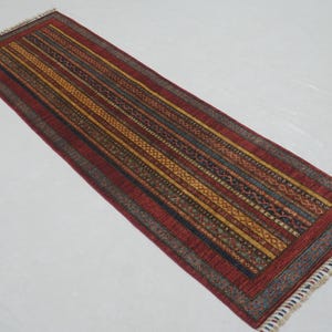 Może przedstawiać: Prostokątny dywanik z czerwonym polem i wzorem w paski w odcieniach ż&oacute;łci, brązu i niebieskiego. Dywan ma frędzelkową krawędź i jest prawdopodobnie wykonany z tkanego materiału.