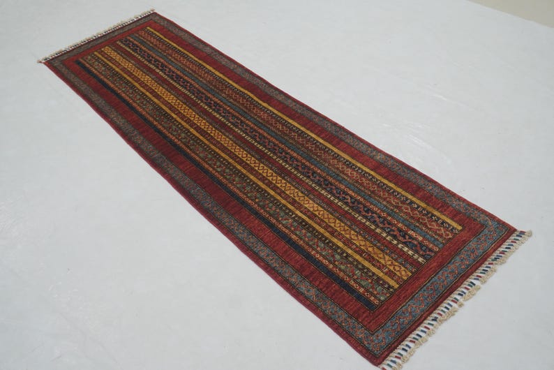 Może przedstawiać: Prostokątny dywanik z czerwonym polem i wzorem w paski w odcieniach ż&oacute;łci, brązu i niebieskiego. Dywan ma frędzelkową krawędź i jest prawdopodobnie wykonany z tkanego materiału.