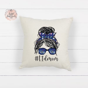 May include: A white throw pillow with a black and white illustration of a woman with a messy bun. The bun is decorated with a blue and white patterned headband. The woman is wearing sunglasses with a blue and white patterned reflection. The text "#T1Dmom" is printed below the illustration.