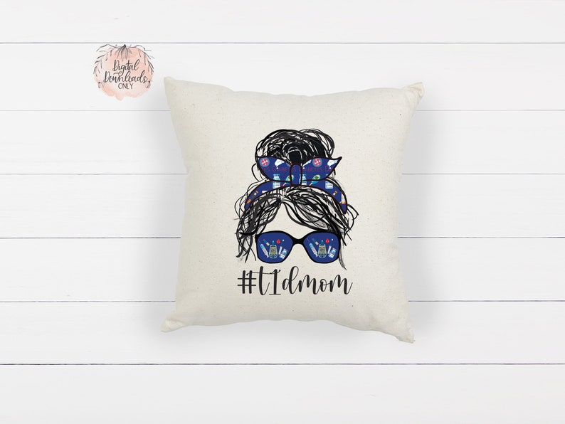 May include: A white throw pillow with a black and white illustration of a woman with a messy bun. The bun is decorated with a blue and white patterned headband. The woman is wearing sunglasses with a blue and white patterned reflection. The text "#T1Dmom" is printed below the illustration.