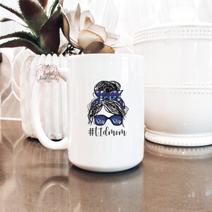 May include: White ceramic mug with a graphic of a woman with a messy bun, sunglasses, and a blue and white patterned headband. The text "#tidmom" is printed below the graphic.