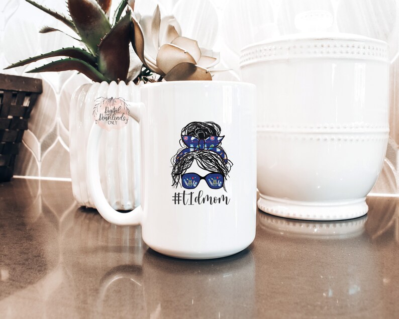 May include: White ceramic mug with a graphic of a woman with a messy bun, sunglasses, and a blue and white patterned headband. The text "#tidmom" is printed below the graphic.