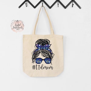 May include: A beige tote bag with a black and white illustration of a woman with a messy bun and sunglasses. The woman's hair is tied with a blue and white patterned ribbon. The text "#T1DMOM" is printed below the illustration.