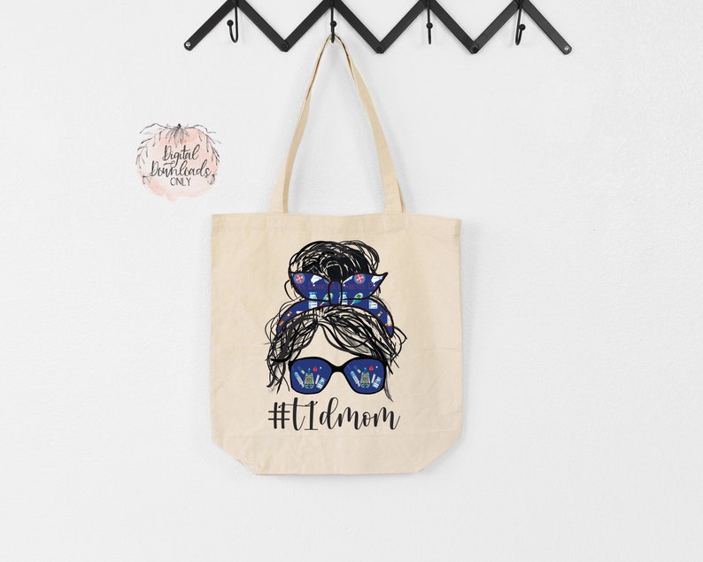 May include: A beige tote bag with a black and white illustration of a woman with a messy bun and sunglasses. The woman's hair is tied with a blue and white patterned ribbon. The text "#T1DMOM" is printed below the illustration.