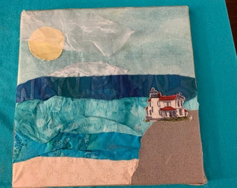 May include: A fabric art piece depicting a white lighthouse with a red roof on a cliff overlooking a blue ocean with a yellow sun in the sky.