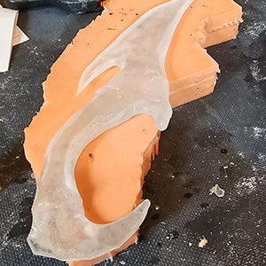 Może przedstawiać: Przezroczysty plastikowy odlew o zakrzywionym kształcie, spoczywający na jasno pomarańczowym silikonowym odlewie. Silikonowy odlew znajduje się na czarnej, teksturowanej powierzchni.