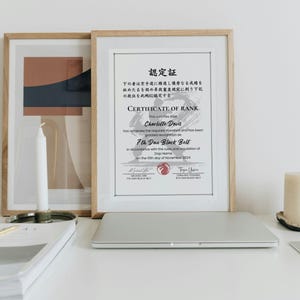 May include: A framed certificate of rank, with Japanese text and English text, is displayed on a white surface. The certificate is for a 7th Dan Black Belt. A laptop, candles, and a stack of papers are also on the surface.