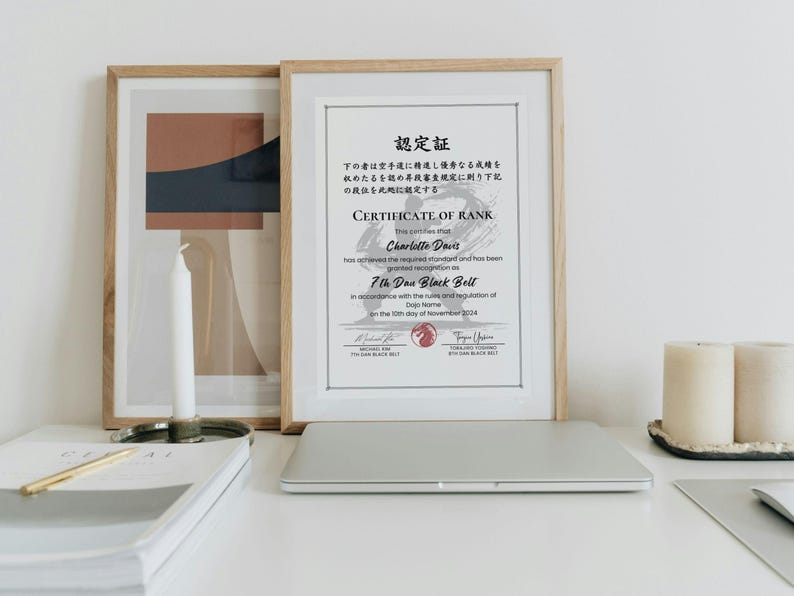 May include: A framed certificate of rank, with Japanese text and English text, is displayed on a white surface. The certificate is for a 7th Dan Black Belt. A laptop, candles, and a stack of papers are also on the surface.
