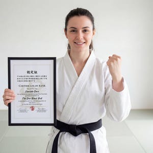 May include: A woman in a white karate uniform with a black belt holds a framed certificate. The certificate, in English and Japanese, is for 7th Dan Black Belt. She smiles and clenches her fist in a gesture of triumph.