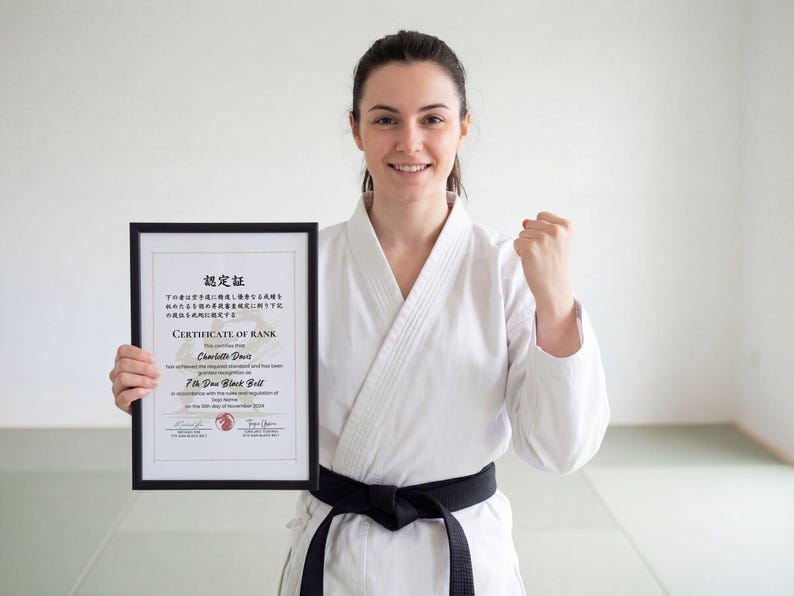 May include: A woman in a white karate uniform with a black belt holds a framed certificate. The certificate, in English and Japanese, is for 7th Dan Black Belt. She smiles and clenches her fist in a gesture of triumph.