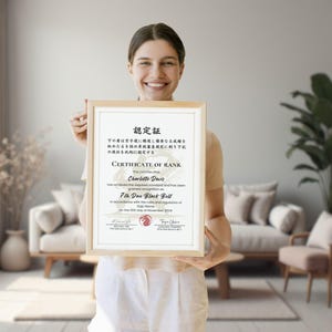May include: A framed certificate with a light wood frame is held up. The certificate has text in English and Japanese. The text reads "Certificate of Rank" and "7th Dan Black Belt". The background shows a living room with a couch and plants.