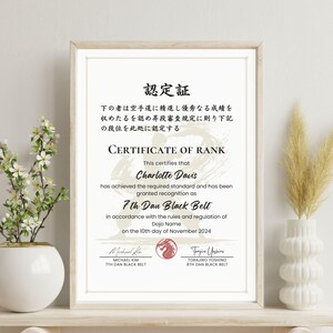 May include: Framed certificate of rank for Charlotte Davis, 7th Dan Black Belt, dated November 10, 2024. The certificate is in a wooden frame and includes Japanese text. A white vase with flowers and a decorative vase with dried plants are on either side.