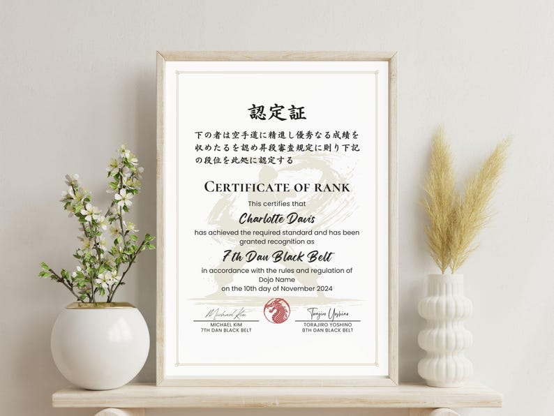 May include: Framed certificate of rank for Charlotte Davis, 7th Dan Black Belt, dated November 10, 2024. The certificate is in a wooden frame and includes Japanese text. A white vase with flowers and a decorative vase with dried plants are on either side.
