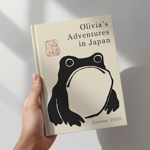Peut inclure: Un livre crème intitulé "Olivia's Adventures in Japan" avec une illustration de grenouille noire. Il présente des caractères japonais rouges et le texte "Summer 2026".
