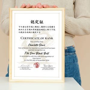 May include: A framed certificate with a light wood frame. The certificate reads "CERTIFICATE OF RANK" and certifies Charlotte Davis as a 7th Dan Black Belt. The date on the certificate is November 10th, 2024.