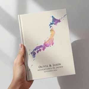 May include: A cream-colored book with a watercolor map of Japan in shades of purple, blue, and yellow. The text "OLIVIA & JASON ADVENTURES IN JAPAN SEPTEMBER 2026" is printed on the cover.