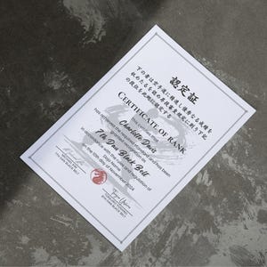 May include: A white certificate with Japanese text and English text, including "CERTIFICATE OF RANK" and "7th Dan Black Belt." The certificate is awarded to Charlotte Davis and dated November 10, 2024. The certificate is on a dark gray surface.