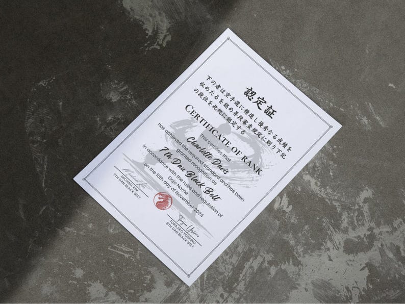 May include: A white certificate with Japanese text and English text, including "CERTIFICATE OF RANK" and "7th Dan Black Belt." The certificate is awarded to Charlotte Davis and dated November 10, 2024. The certificate is on a dark gray surface.