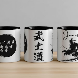 May include: Three white ceramic mugs with black rims. The mug on the left has black calligraphy in a circle. The mug in the middle has black calligraphy that reads "Bushido". The mug on the right has a black silhouette of a samurai warrior with the name "James" written above it.