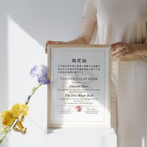 May include: A framed certificate with Japanese text and English text, held by a person. The certificate is for a 7th Dan Black Belt, with the name Charlotte Davis. The frame is light wood, and the background is white.