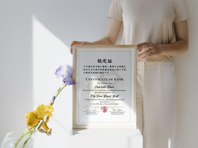 May include: A framed certificate with Japanese text and English text, held by a person. The certificate is for a 7th Dan Black Belt, with the name Charlotte Davis. The frame is light wood, and the background is white.
