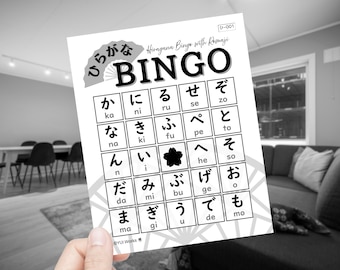 May include: A black and white Hiragana Bingo card with Romaji. The card features a grid of Japanese characters and the word "BINGO" in large letters. A hand holds the card in front of a blurred interior.