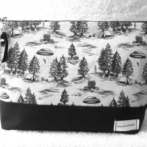 May include: Black and white zippered pouch with a print of trees, tents, and UFOs. The pouch has a black bottom and a white top with a black zipper.