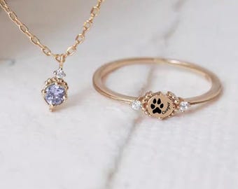May include: A gold-toned necklace with a small, round, light purple stone pendant and a matching gold-toned ring featuring a central circular disc engraved with a black paw print and the name Cooper, flanked by two tiny clear stones on each side.