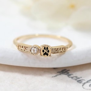 Puede incluir: Un anillo dorado con una huella de pata negra y una piedra transparente. El anillo tiene un diseño ornamentado en la banda. El anillo está sobre una superficie blanca.