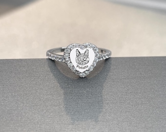 Könnte beinhalten: Ein silberner Ring mit herzförmiger Fassung, der eine Katzenillustration und den Namen "Potato" zeigt. Der Ring ist mit kleinen, klaren Steinen um das Herz und entlang des Bandes besetzt, was einen funkelnden Effekt erzeugt.