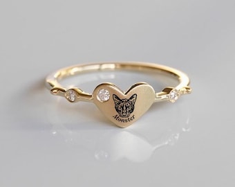 Könnte beinhalten: Goldfarbener Ring mit herzförmigem Mittelstück. Das Herz zeigt ein schwarzes Katzen-Gesicht und das Wort "Monster". Kleine klare Steine sind auf beiden Seiten des Herzens angebracht. Ein zartes und einzigartiges Schmuckstück.