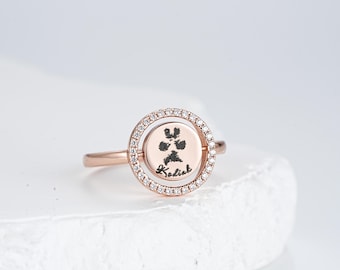 Könnte beinhalten: Ein roségoldfarbener Ring mit einer drehbaren runden Scheibe in der Mitte, die mit einem schwarzen Pfotenabdruck und dem Namen Kodiak graviert ist. Der äußere Rand des kreisförmigen Rahmens ist mit kleinen klaren Steinen besetzt.