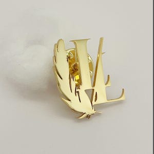 May include: A gold-colored pin featuring the letters "LV" and a feather design. The pin is made of shiny metal and has a classic, elegant style. The pin is likely a fashion accessory.