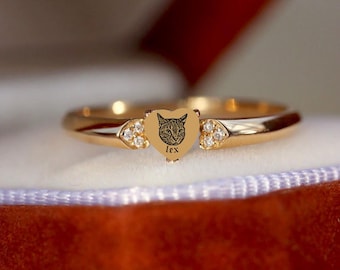 May include: A gold-toned ring featuring a central heart-shaped charm engraved with a cat face and the name lex. The charm is flanked by two small triangular accents, each set with three clear, sparkling stones on the polished metal band.