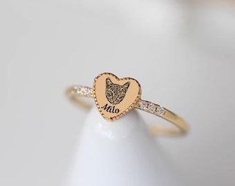 Könnte beinhalten: Goldfarbener Ring mit herzförmiger Fassung. Die Fassung zeigt eine Gravur eines Katzenkopfes und den Schriftzug "Milo". Der Ring ist mit kleinen, klaren Steinen besetzt.