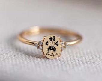 Könnte beinhalten: Goldfarbener Ring mit einem ovalen Mittelteil, das einen Pfotenabdruck und das Wort "Stripes" zeigt. Der Ring ist mit kleinen, klaren Edelsteinen auf beiden Seiten des Mittelteils verziert. Ein zartes und elegantes Schmuckstück.