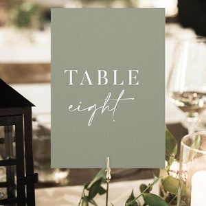 May include: A sage green table number card with white text that reads "TABLE eight". The card is on a metal stand with greenery.