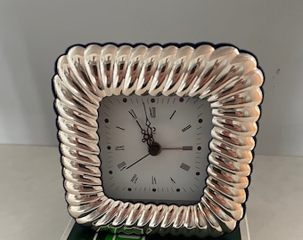 May include: A silver square clock with Roman numeral markings on a white face. The clock is in a silver frame with a decorative, rope-like design. The clock is sitting on a green and white box with the text "MADE IN ITALY" and "336 FI" printed on it.