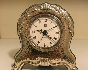 May include: A silver-toned, ornate, quartz clock with Roman numeral markings on the face. The clock has a hammered metal finish and sits on a decorative base.
