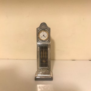 May include: A silver-colored miniature grandfather clock with a gold-colored clock face. The clock has a rectangular body with decorative scrollwork and a dark brown panel. The clock face displays the time.