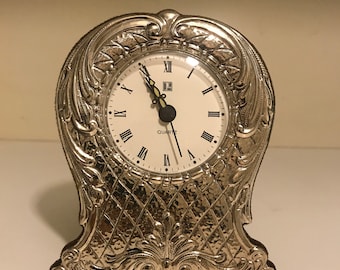 May include: Silver-toned table clock with an ornate design and a white face with black Roman numeral markings. The clock face reads "QUARTZ".