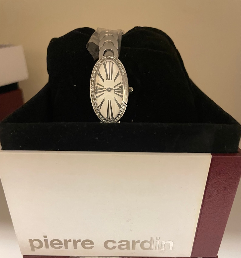 K&ouml;nnte beinhalten: Eine silberne ovale Uhr mit wei&szlig;em Zifferblatt und r&ouml;mischen Ziffern f&uuml;r die Stunden. Die Uhr befindet sich in einer schwarzen Samtbox mit dem Aufdruck "pierre cardin" auf der Vorderseite.