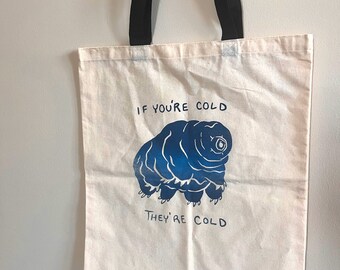 Puede incluir: Una bolsa de lona blanca con correas negras. La bolsa tiene una ilustración azul de un tardígrado con el texto "If you're cold they're cold".