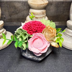 May include: A floral arrangement featuring a variety of colorful wooden flowers in a dark gray geometric vase. The flowers include a vibrant red peony, a light pink rose, and a tan-colored flower, with green leaves and stems.