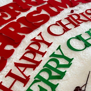 May include: Red and green felt letters spelling "MERRY CHRISTMAS" and "HAPPY CHRISTMAS" on a white, textured surface. Decorative sprigs with berries and silver accents are also present, suggesting holiday decor.