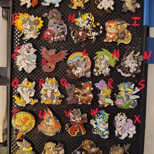 May include: A collection of twenty-eight colorful enamel pins featuring various anthropomorphic animal characters, each labeled with a red letter or number, arranged in a grid on a black perforated display board.